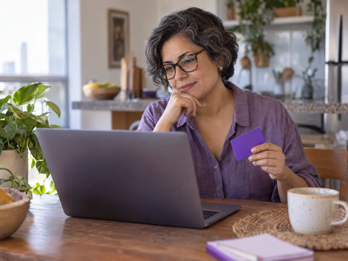 Mulher na mesa da cozinha segura um cartão ao lado do notebook
